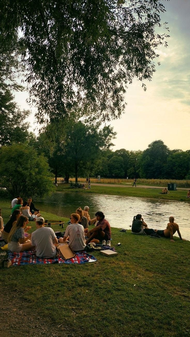 many people are sitting on the grass next to a river and picnicking at sunset