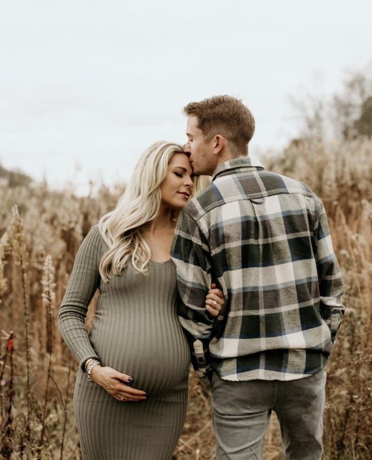 a pregnant couple standing next to each other in front of some tall grass and trees