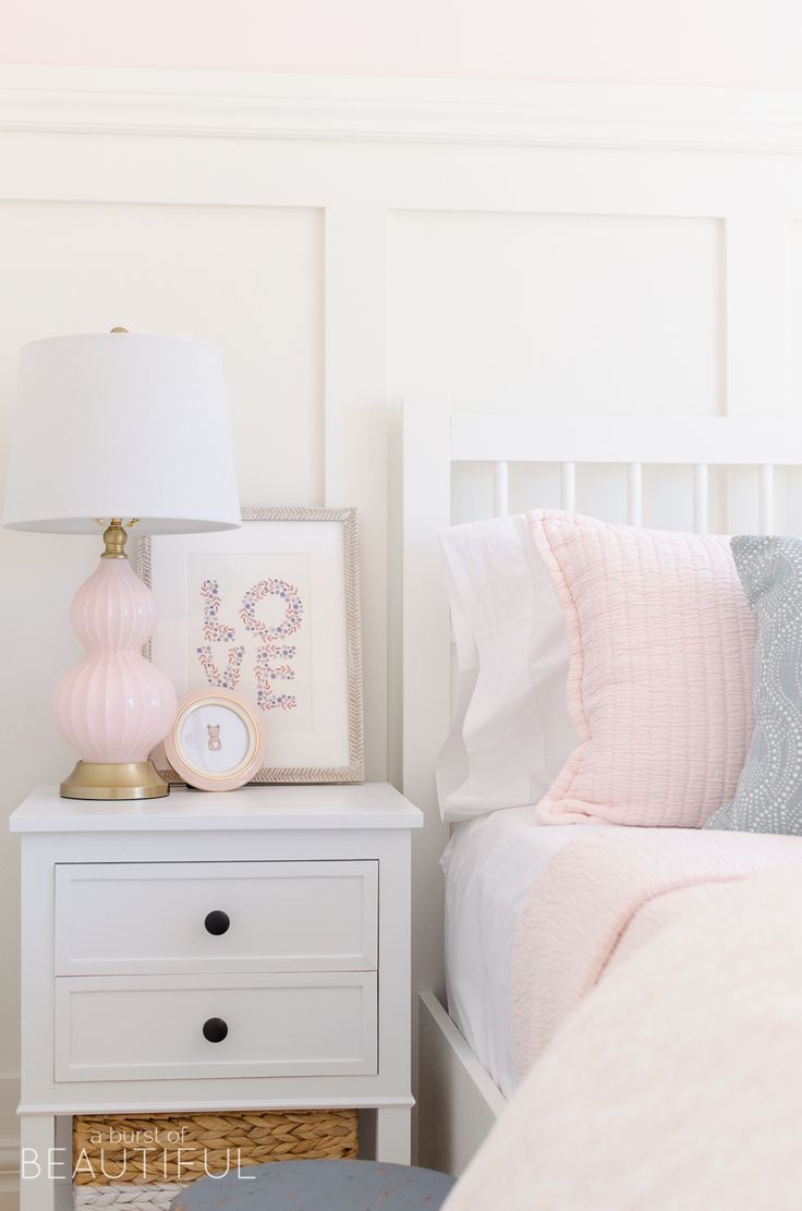 a white bed with pink and blue decor on it's night stand next to a nightstand