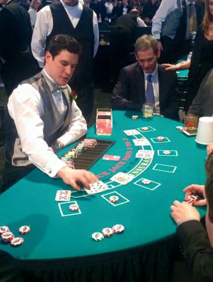 two men playing cards at a casino table with other people around the tables looking on