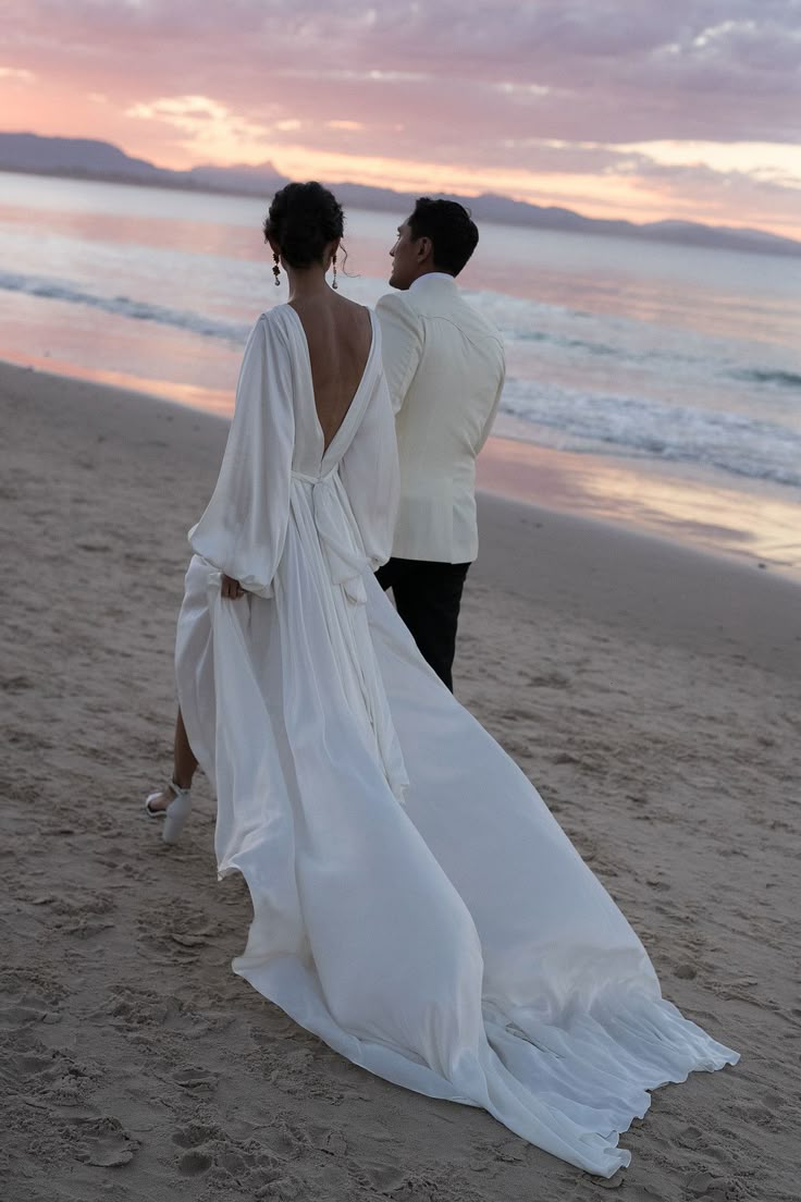 a bride and groom walking on the beach at sunset with their back to the camera