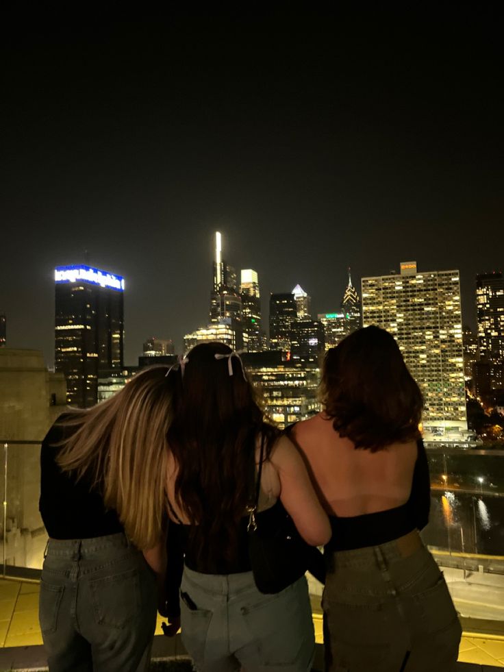 three women standing on top of a roof looking at the city lights in the distance