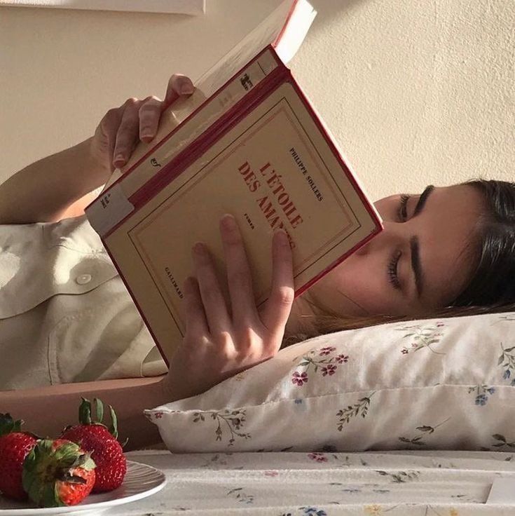 a woman laying in bed reading a book with strawberries on the table next to her