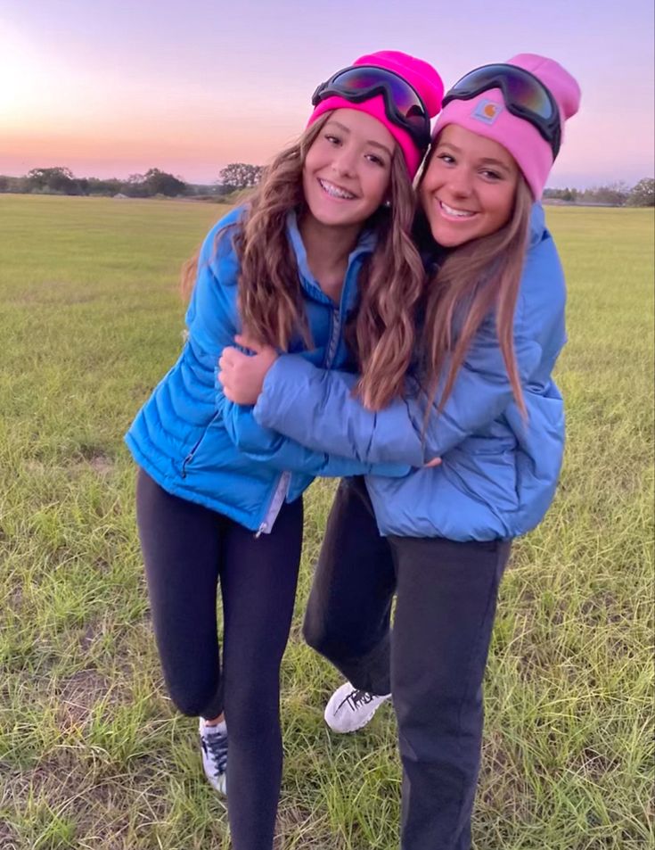 two girls are hugging in the middle of an open field at sunset with their ski goggles on