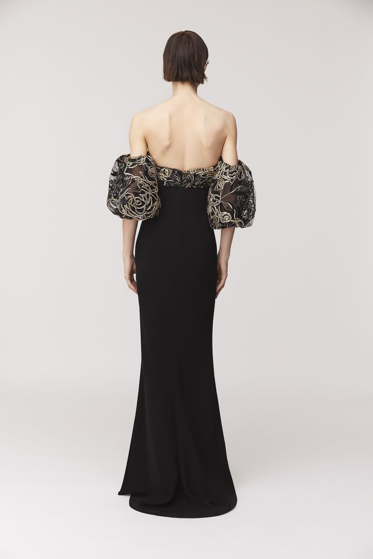 the back of a woman in a black dress with gold embroidered sleeves and an open shoulder