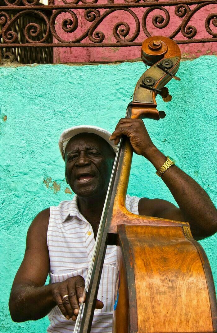 a man with a hat and glasses holding a cello in front of a blue wall