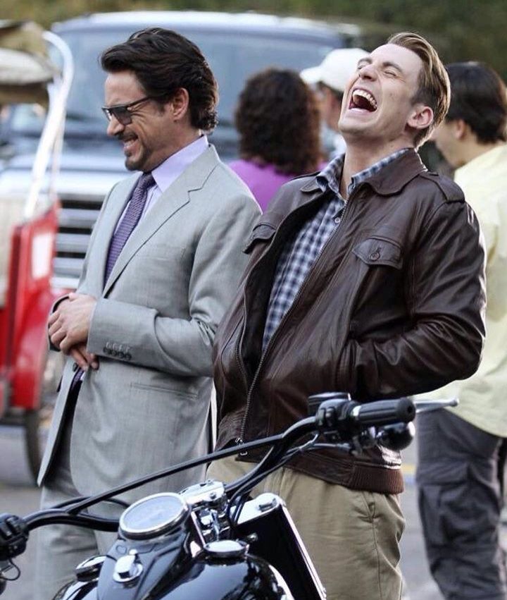 two men laughing while standing next to a motorcycle