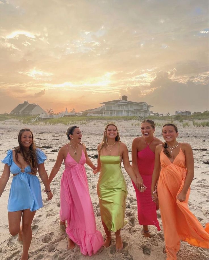 four women in dresses are walking on the beach at sunset with one holding her hand out