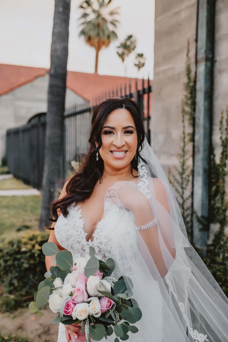 a woman in a wedding dress holding a bouquet and smiling at the camera with palm trees behind her