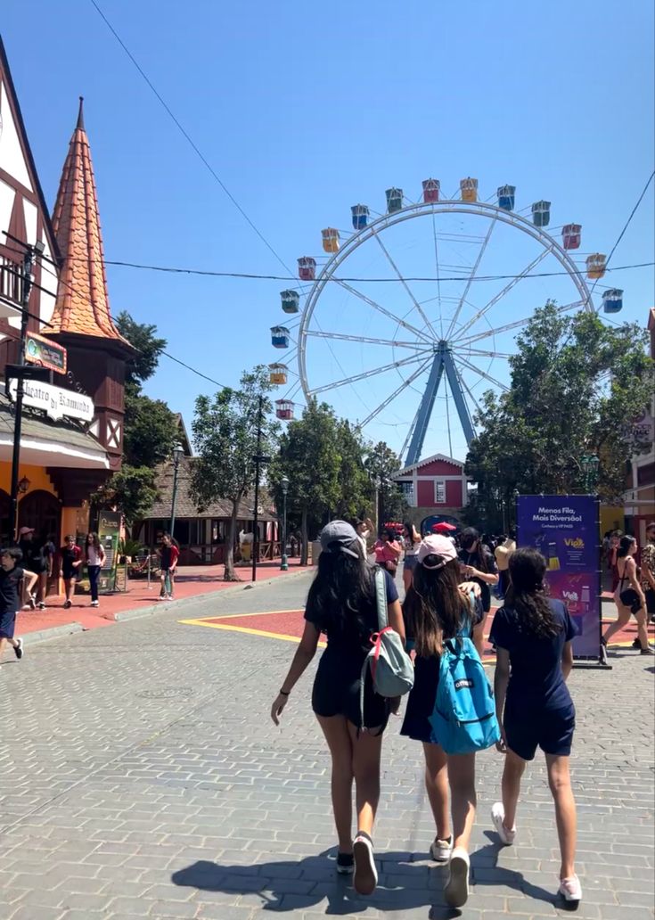 three girls are walking down the street in front of an amusement park ride and ferris wheel