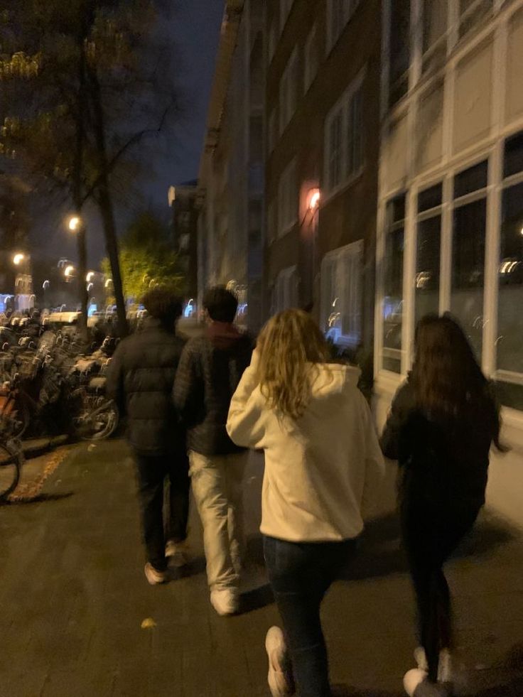 a group of people walking down a sidewalk next to a building at night with bikes parked on the street