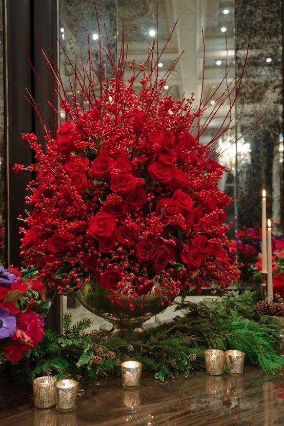 red flowers and greenery are arranged in a vase on the table next to candles