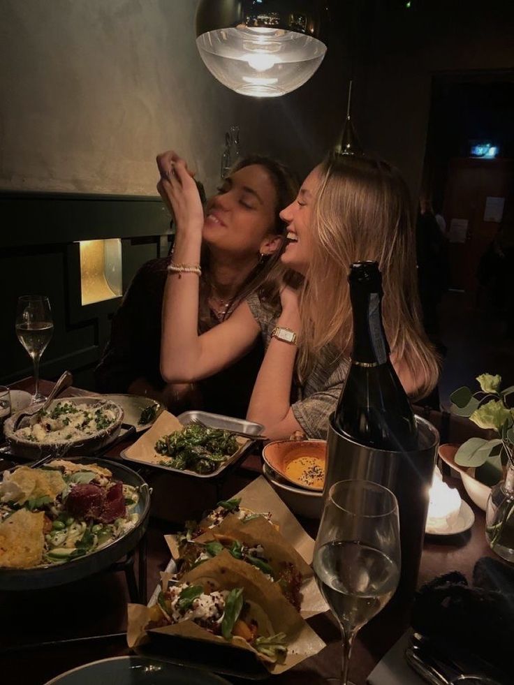 two women sitting at a table with food and drinks in front of them, one kissing the other