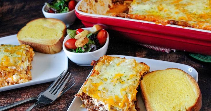 two plates with lasagna sandwiches on them next to a bowl of salad and bread