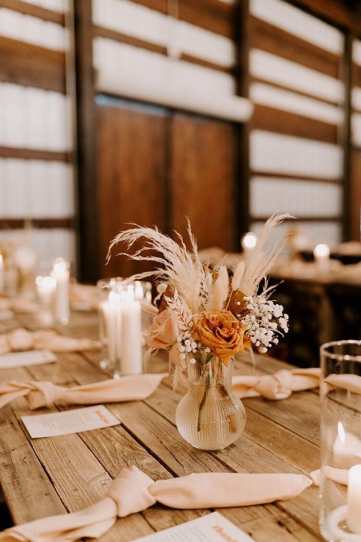 a wooden table topped with lots of white and orange flowers next to tall glass vases filled with candles