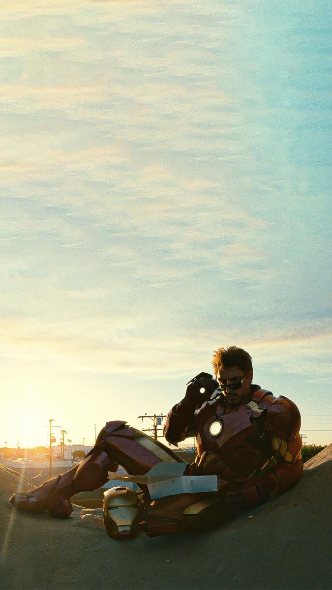 a man dressed as iron man sitting on the ground talking on a cell phone and holding his hand up to his ear