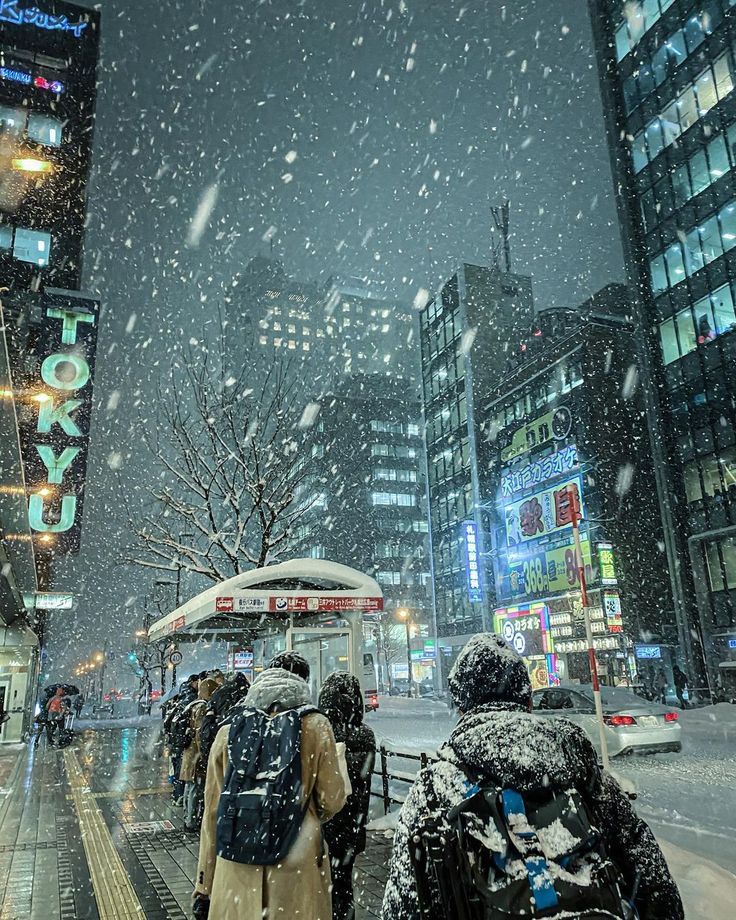 people are walking in the snow on a city street with buildings and lights behind them