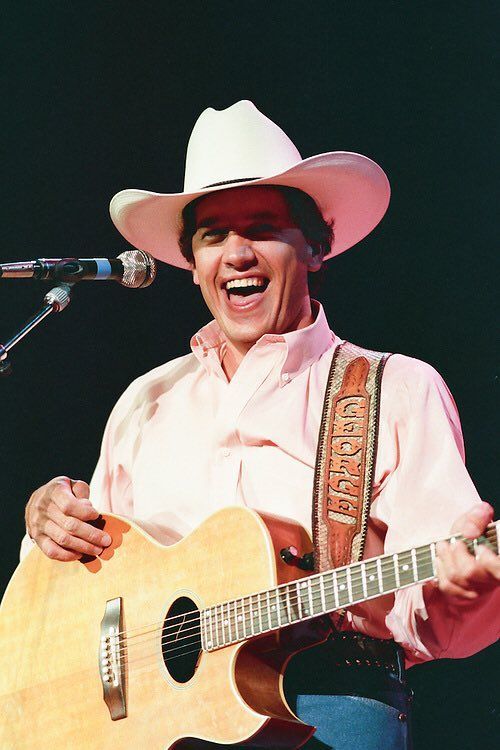 a man wearing a cowboy hat and holding a guitar in front of a microphone on stage