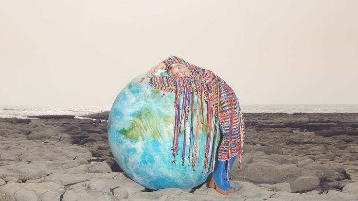 a woman standing on top of a rock covered beach next to the ocean holding a giant blue ball