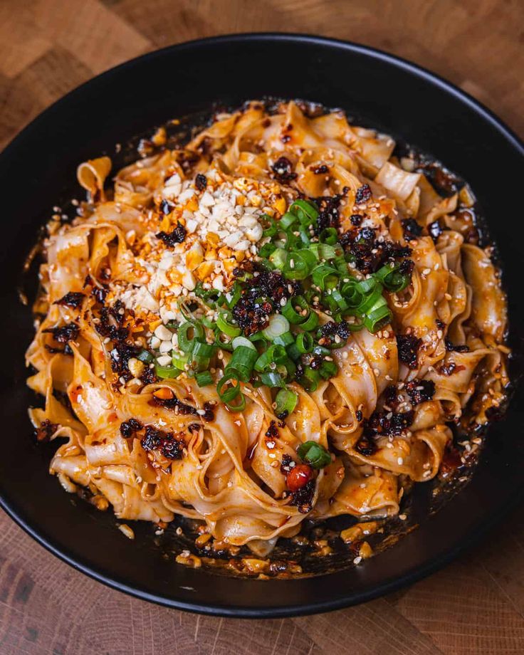 a black bowl filled with noodles and vegetables on top of a wooden table next to a fork