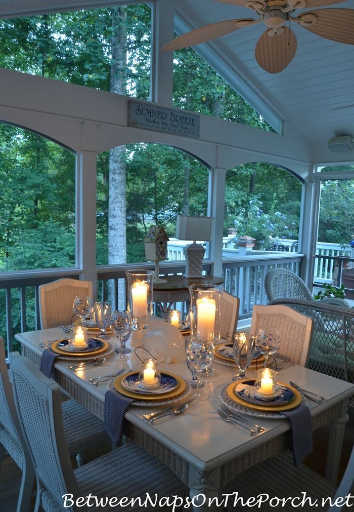 a dining table set with place settings and lit candles on the dinner table in front of an open porch