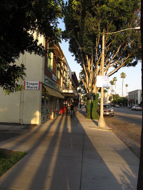 people are walking down the sidewalk in front of shops on a sunny day with long shadows