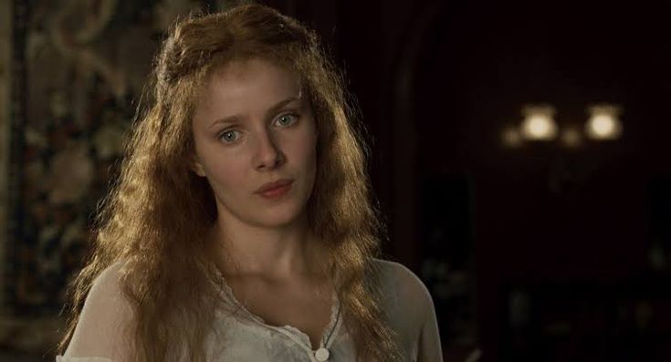 a woman with long red hair in a white shirt looking at the camera while standing in a dark room