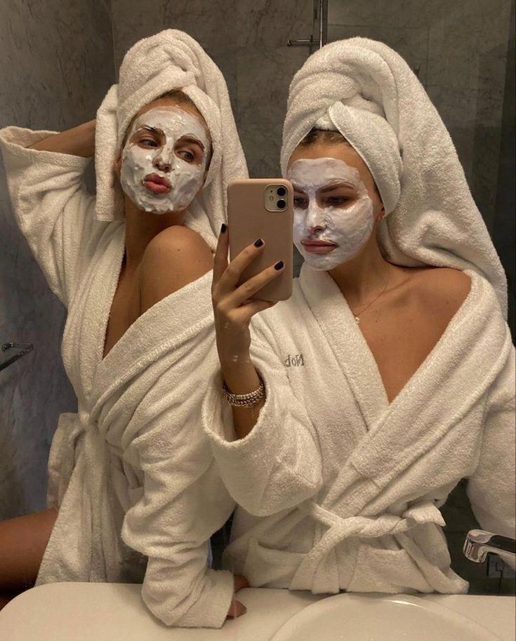 two women with face masks on taking a selfie in the bathroom while wearing white towels