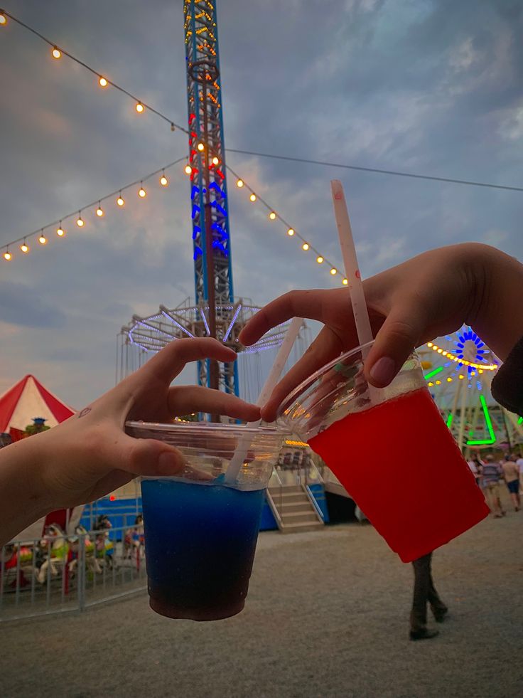 two people holding cups with drinks in front of a carnival ride at night, one is blue and the other has red