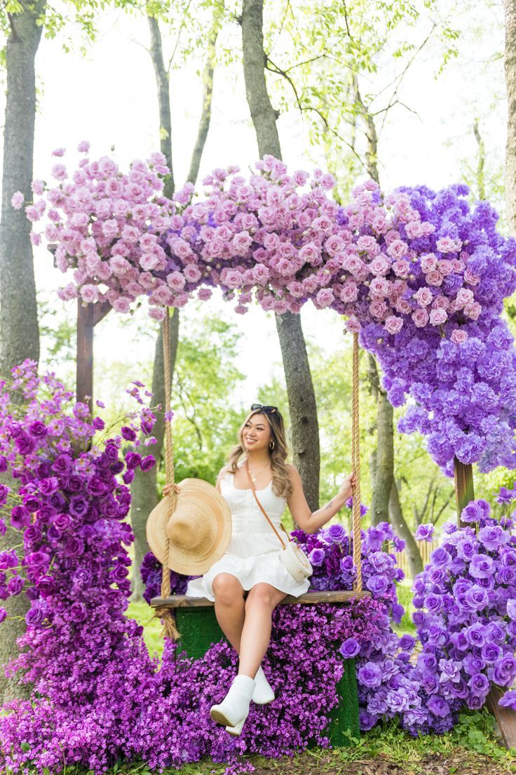 a woman sitting on a swing surrounded by purple flowers in the park, posing for a photo