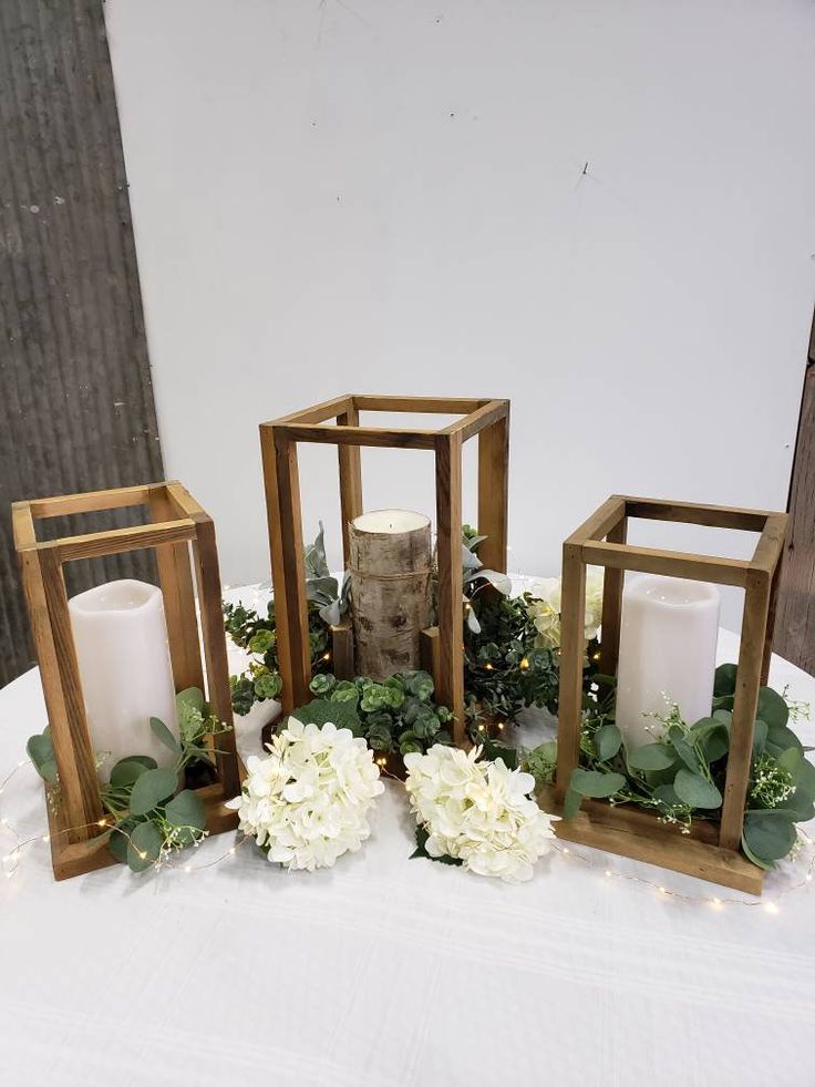 three wooden lanterns with flowers and greenery on a white table cloth covered tablecloth