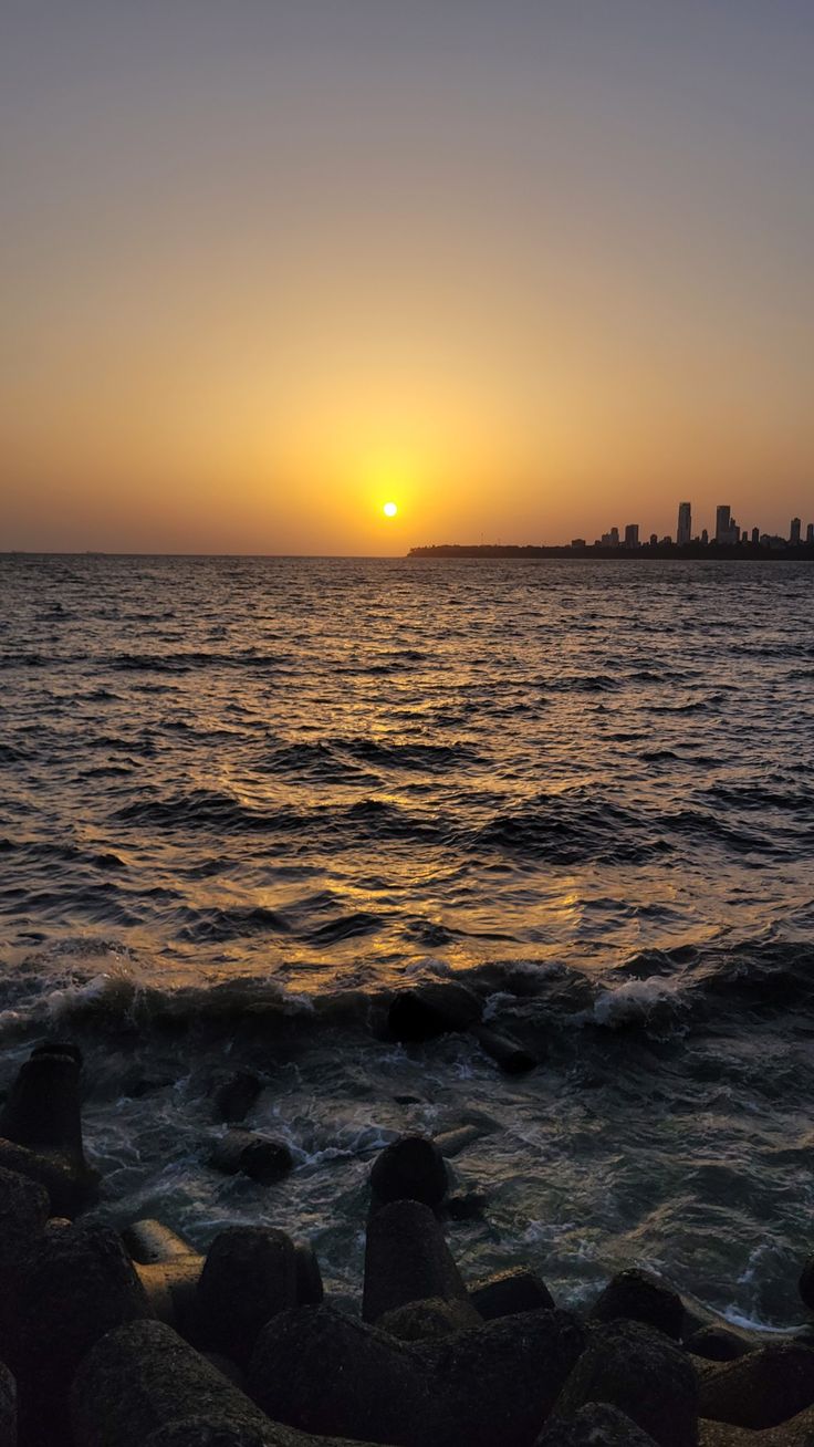 the sun is setting over the ocean with rocks on the shore and buildings in the distance