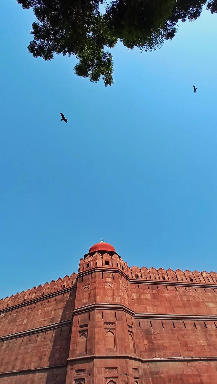 two birds are flying in the blue sky above a large brick building with a red roof