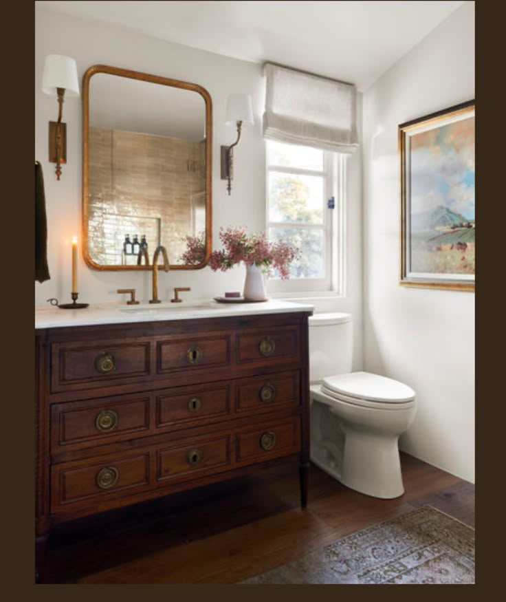 a white toilet sitting next to a wooden dresser in a bathroom under a window with a painting on the wall