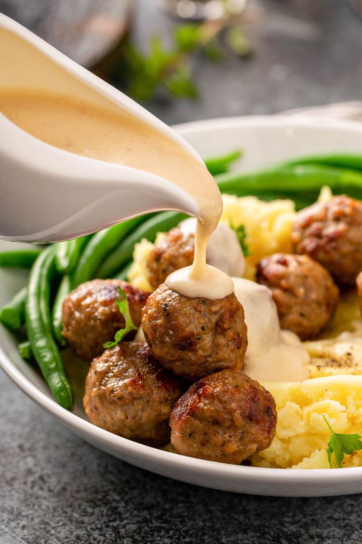 someone pouring sauce on meatballs over mashed potatoes and green beans in a white bowl