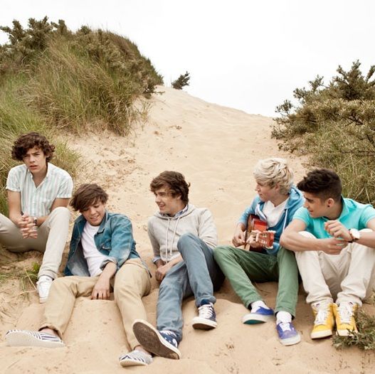 five young men sitting on the sand with their backs to each other and one holding a guitar