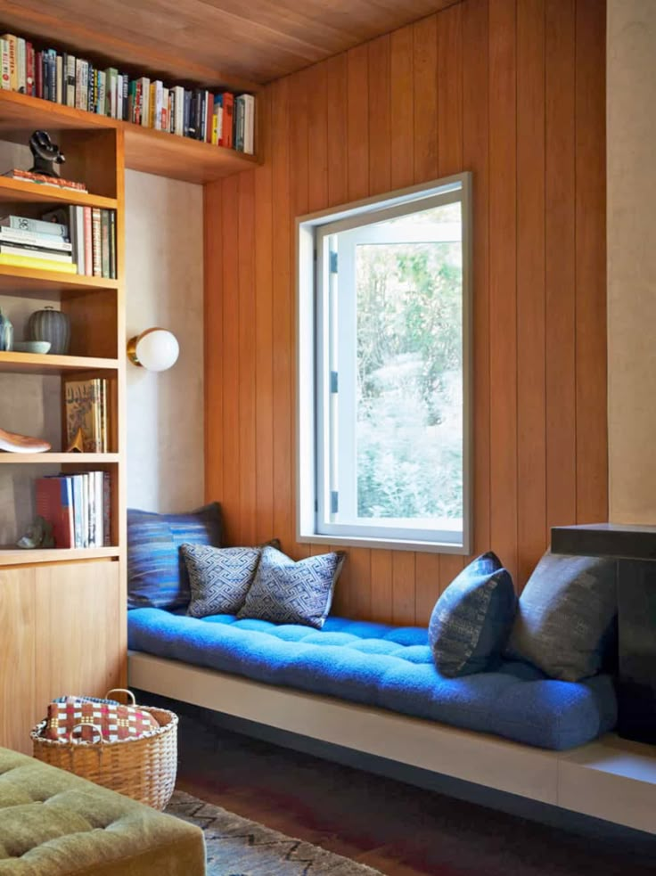 a room with a window, bookshelf and blue cushion on the bench in front of it