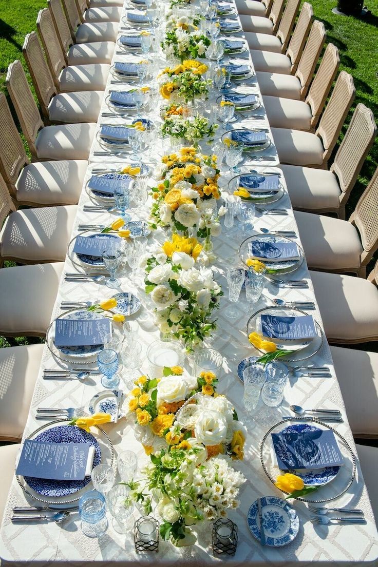 a long table is set with blue and white plates, yellow flowers, and glassware