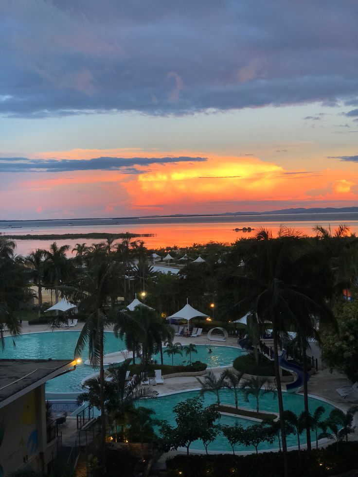 the sun is setting over an outdoor swimming pool with palm trees and beach umbrellas