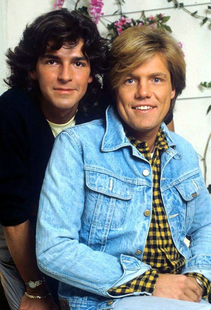 two men sitting next to each other in front of a wall with flowers on it