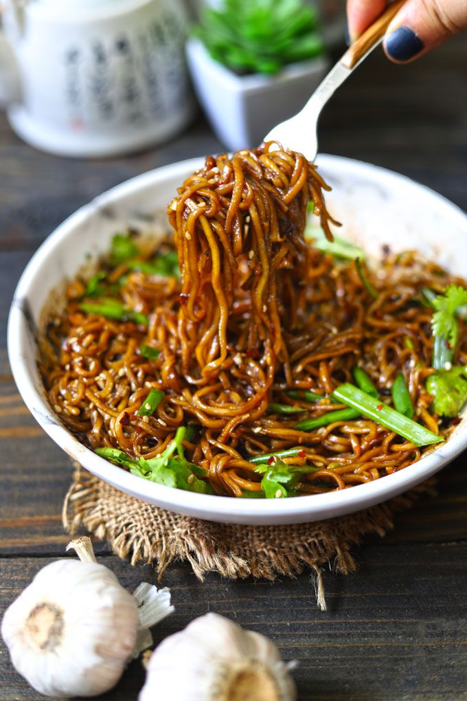 someone is spooning noodles out of a white bowl with green vegetables and garlic on the side