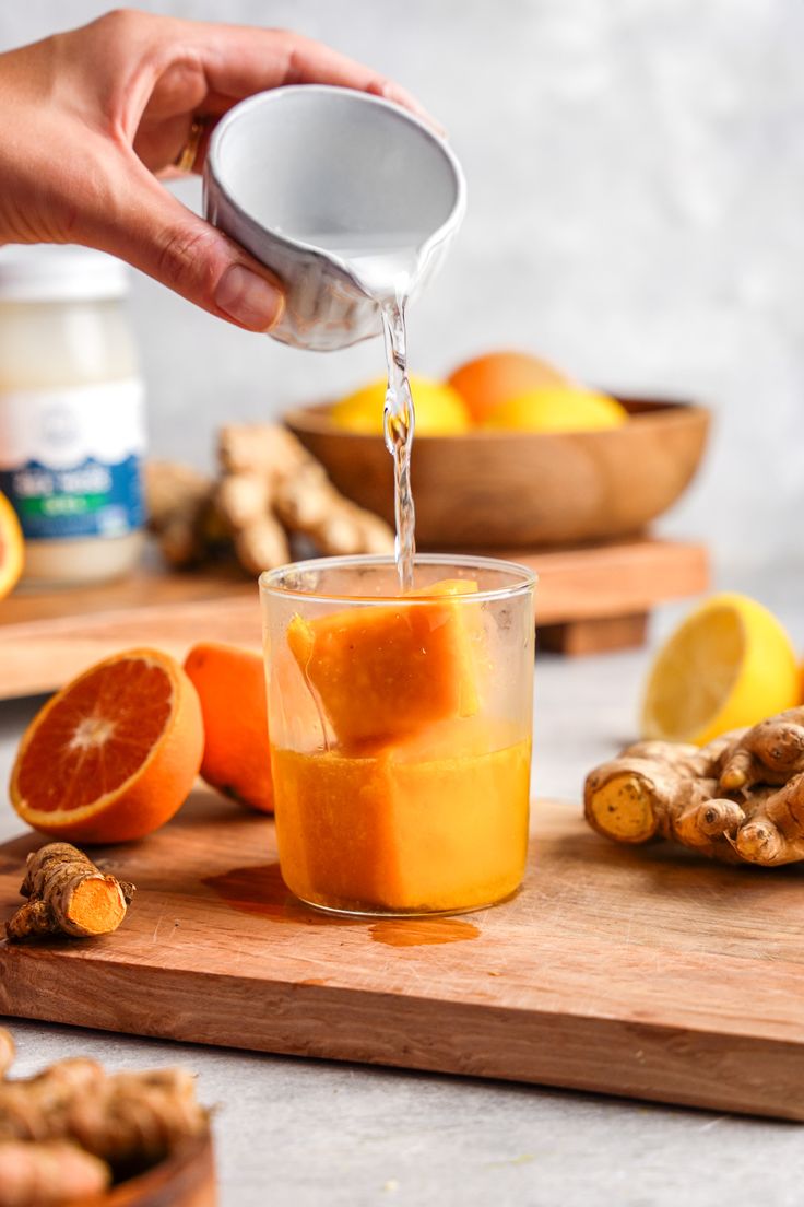 someone pouring orange juice into a glass on top of a cutting board with sliced oranges and ginger