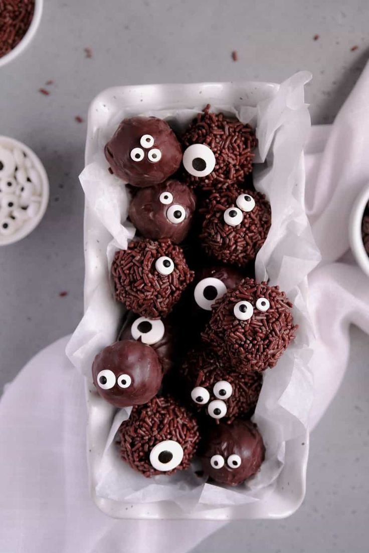 chocolate covered donuts with googly eyes in a white container on a table next to two cups of coffee