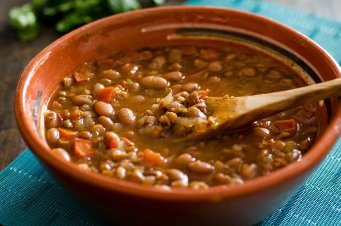 a brown bowl filled with beans and carrots next to a wooden spoon on top of a blue napkin