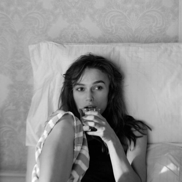 a woman sitting on a bed drinking from a glass in front of her face and looking at the camera