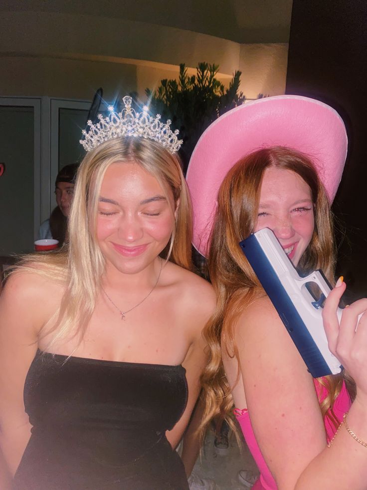 two women in dresses and tiaras smile as they hold up their cell phones to take pictures