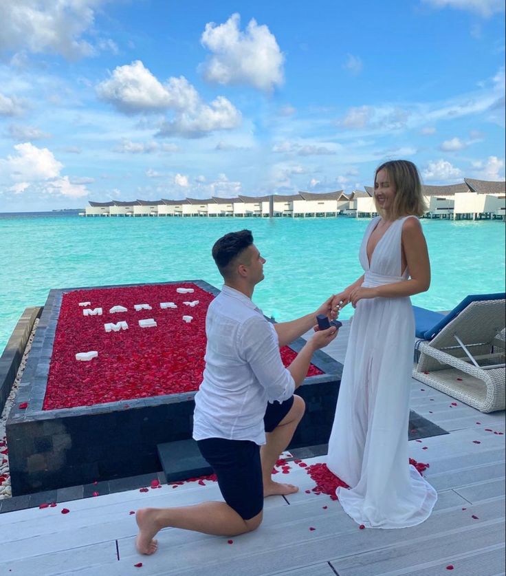 a man kneeling down next to a woman in front of the ocean with red petals on it