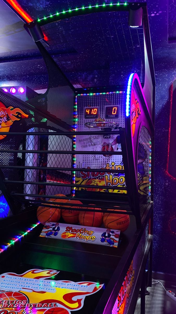 an indoor pinball machine with neon lights and basketballs on the floor in front of it