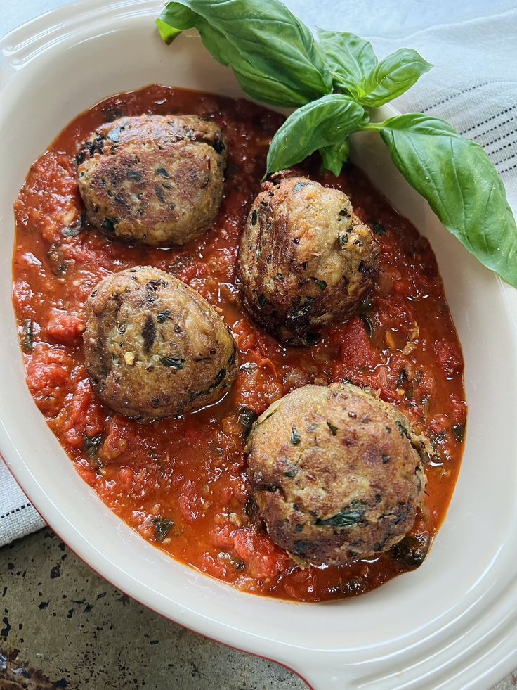 three meatballs in tomato sauce and basil on top of a white plate with a green leafy garnish