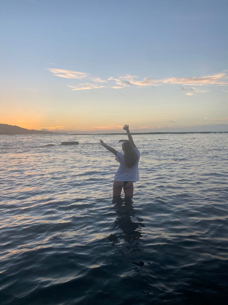 a woman is standing in the water with her arms up to the sky as the sun sets